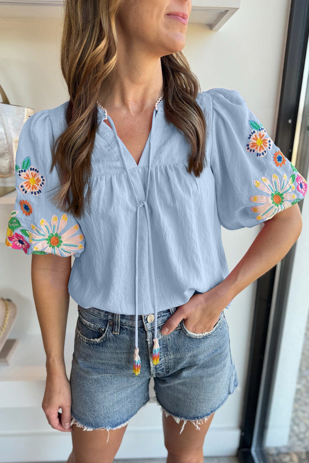 Woman wearing a light blue blouse with colorful floral embroidery and denim shorts indoors.
