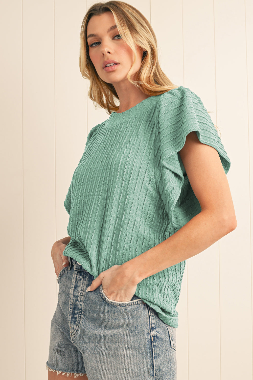 Woman wearing a green textured top and denim shorts against a light background