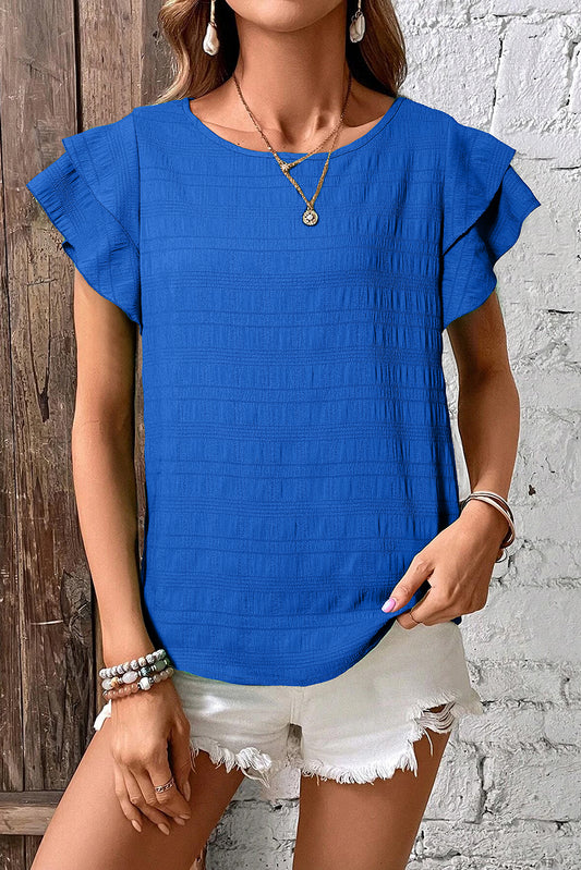 Woman wearing a blue top with ruffled sleeves against a wooden and white brick wall.