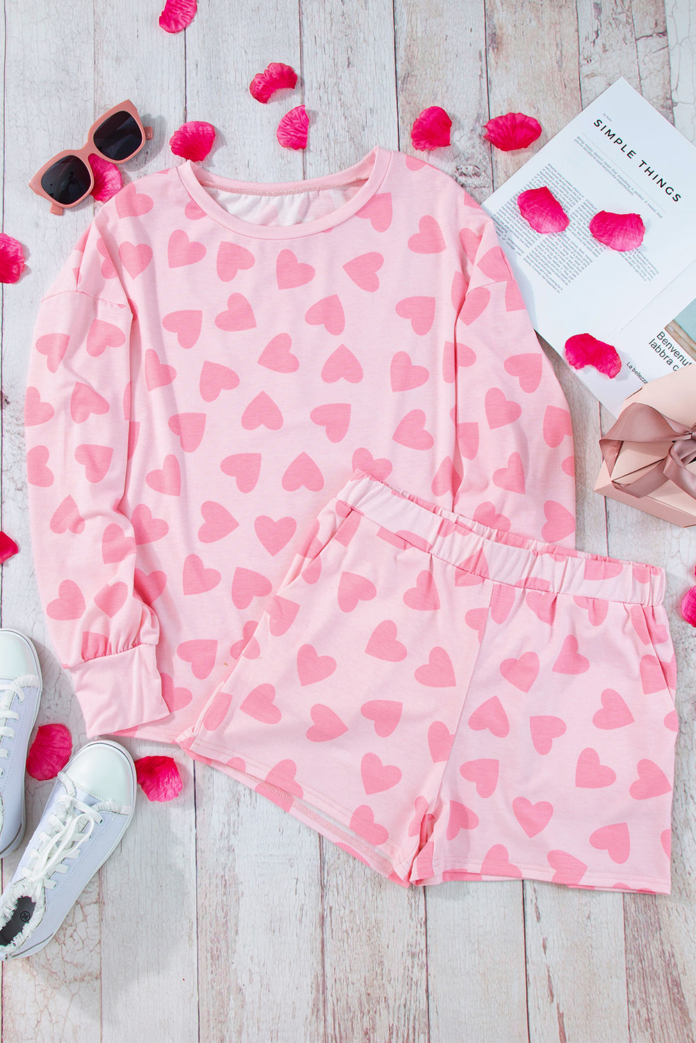 Pink heart-patterned pajama set on a wooden surface with sunglasses and a book.