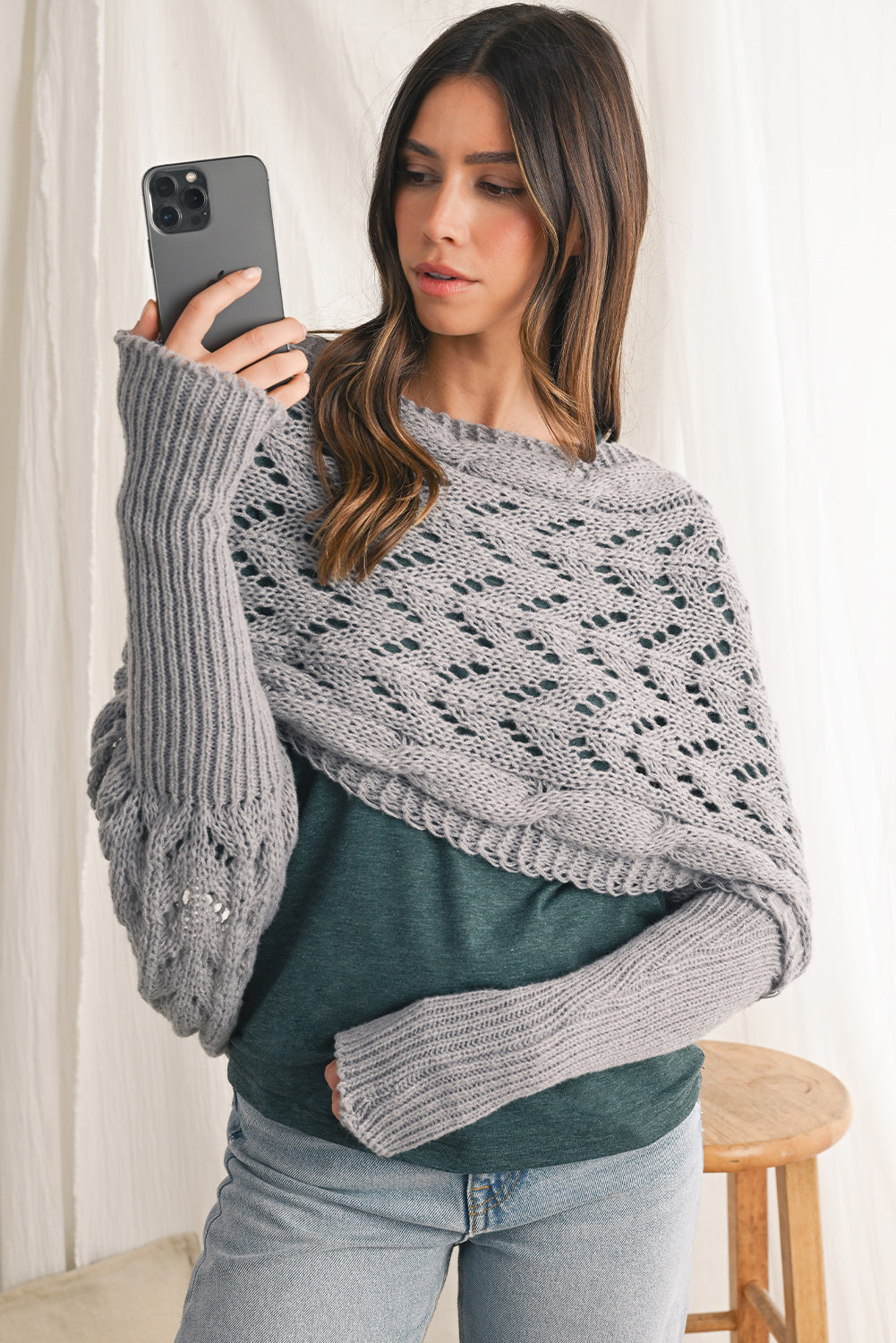 Woman wearing a gray knit poncho over a green sweater, standing indoors.