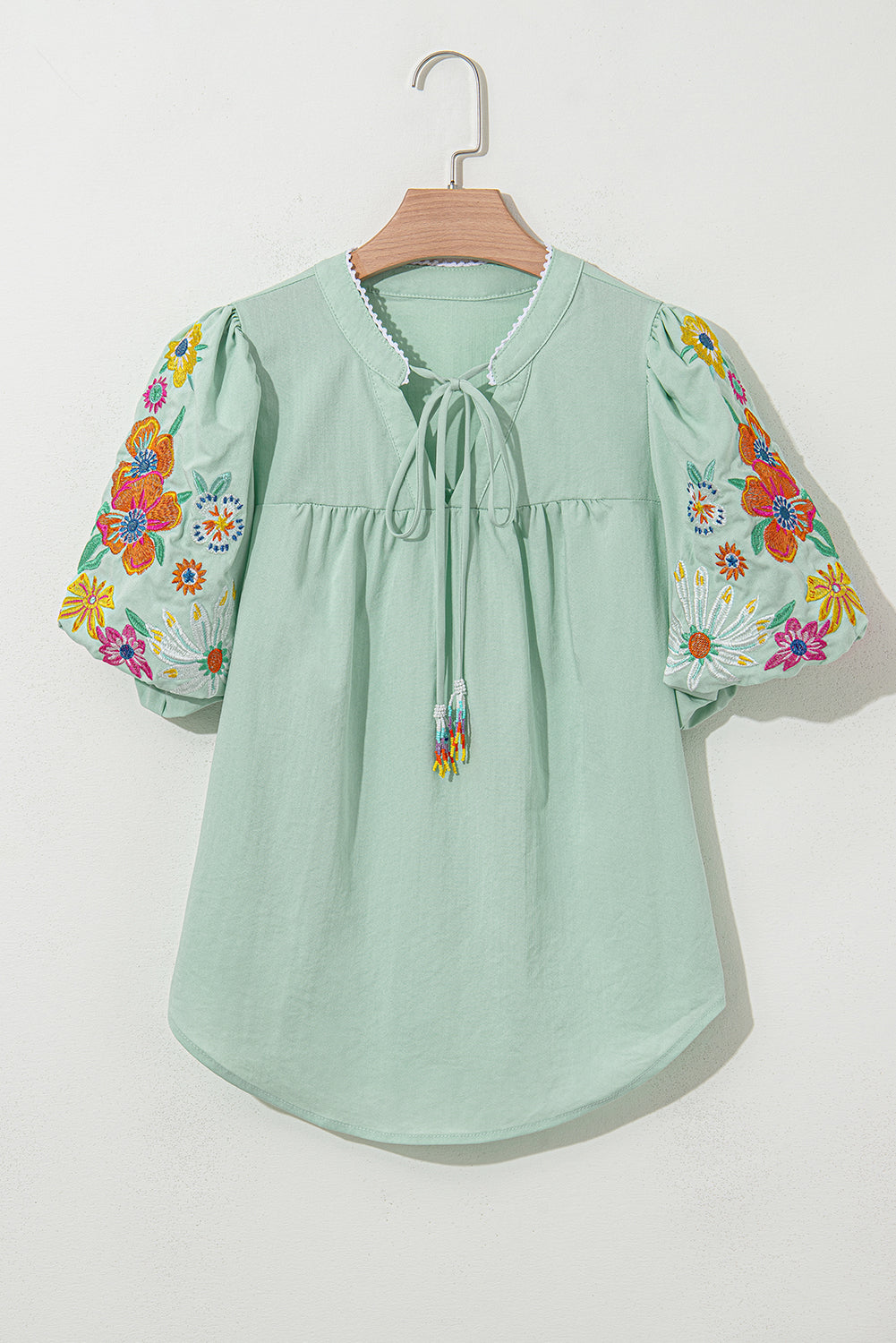 Mint green blouse with floral embroidery on sleeves, hanging on a wooden hanger against a white background.