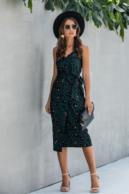 Woman in a dark green dress with a pattern, black hat, and sunglasses standing against a light gray wall.