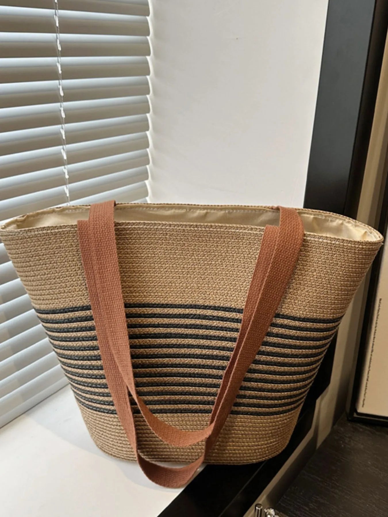 Straw tote bag with brown handles on a surface near a window with blinds.