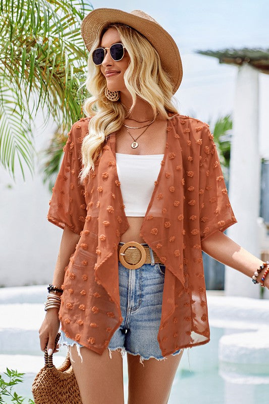 Woman in a rust-colored kimono-style cover-up, white top, and denim shorts standing by a pool.
