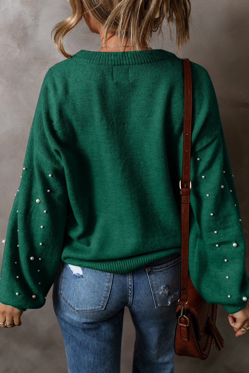 Person wearing a green sweater with pearl embellishments and blue jeans, holding a brown bag.