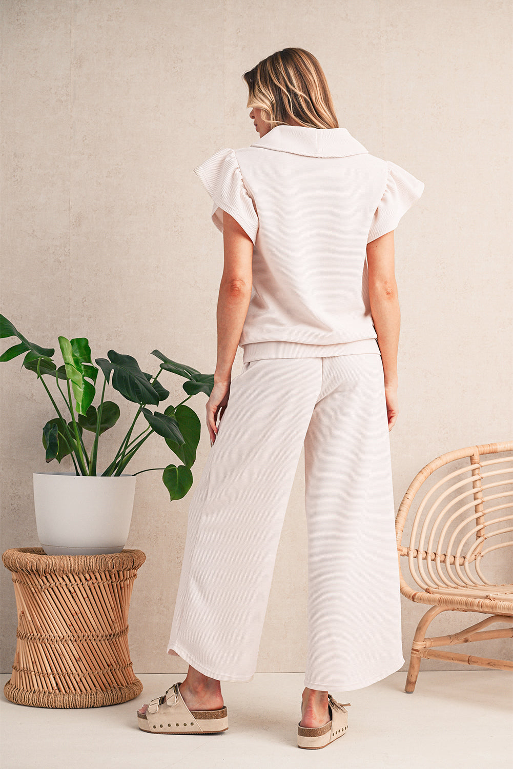 Woman wearing a white outfit standing in a minimalistic room with a plant and chair.