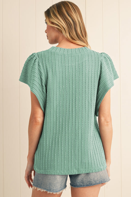 Woman wearing a teal knitted top and denim shorts against a beige wall.