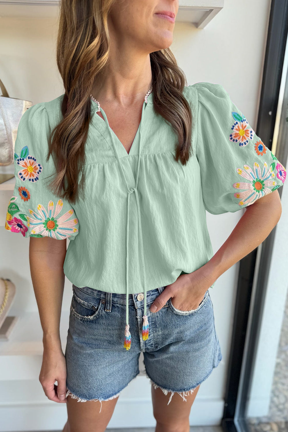 Woman wearing a light green blouse with colorful floral sleeves and denim shorts indoors.