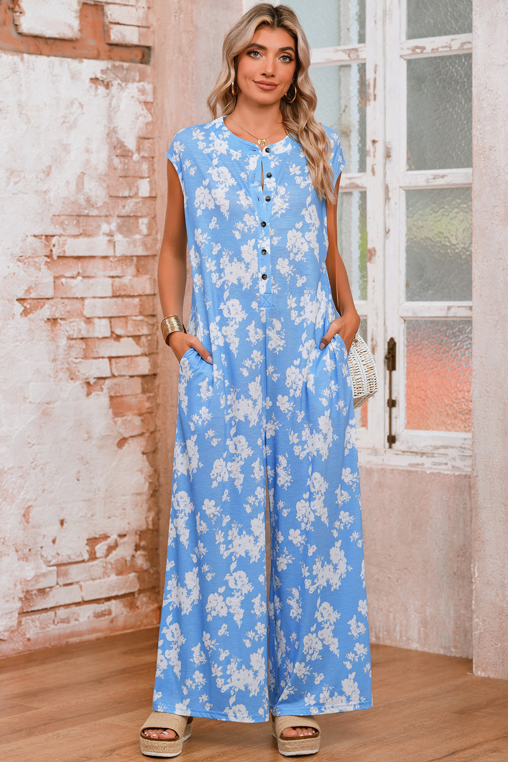 Woman wearing a blue floral jumpsuit standing in a room with a brick wall and window.