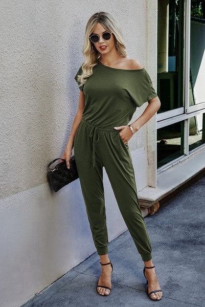 Woman wearing an olive green jumpsuit standing against a light-colored wall.