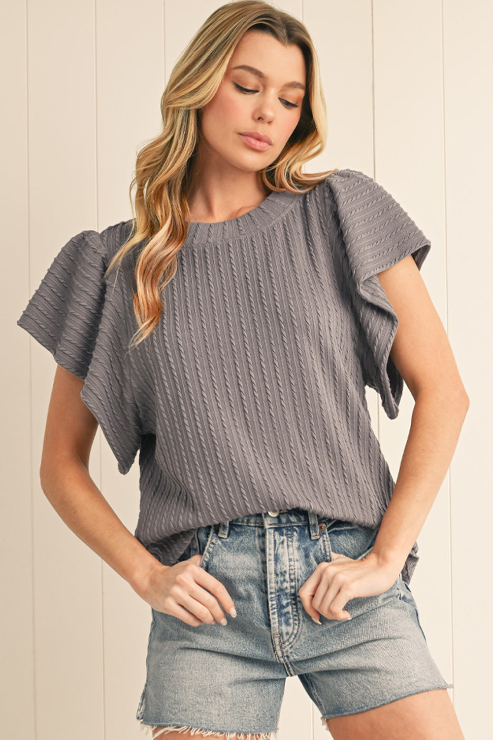 Woman wearing a gray ribbed top and denim shorts against a light wooden panel background