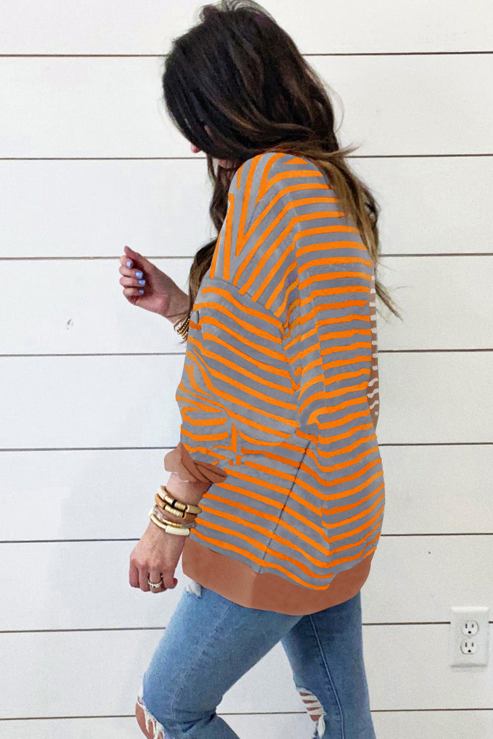 Person wearing a striped orange and beige top with blue jeans against a white wooden panel background.