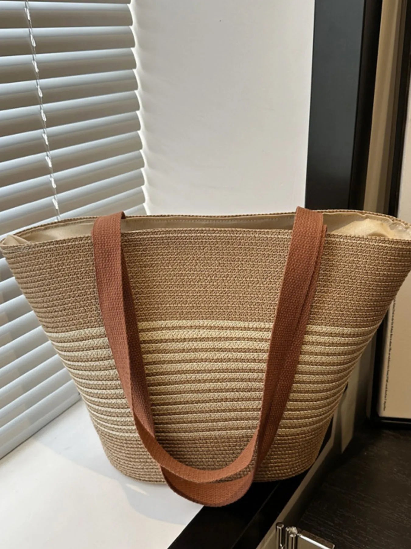 Beige straw tote bag with brown handle on a surface near a window with blinds.