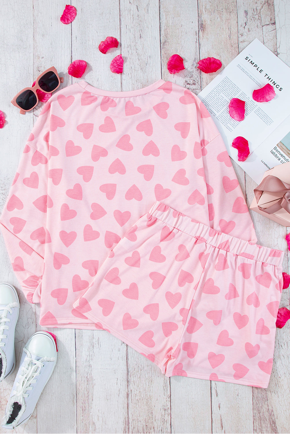 Pink heart-patterned pajama set on a wooden surface with sunglasses and a magazine.